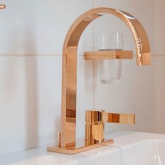 Ein eleganter, gebogener Wasserhahn aus Roségold mit einem Glas und einem Seifenspender im Hintergrund auf einer weißen Fliese.