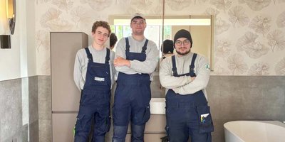 Die drei jungen Männer in Arbeitskleidung posieren selbstbewusst vor einem Spiegel in einem hellen, eleganten Badezimmer.