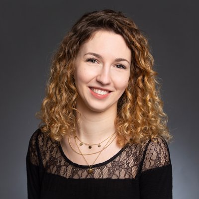 Die Frau hat eine fröhliche Ausstrahlung, ihre lockigen Haare fallen sanft über ihre Schultern und sie trägt mehrere zarte Halsketten.