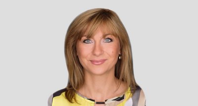 Eine Frau mit blonden Haaren und strahlend blauen Augen lächelt freundlich in die Kamera, gekleidet in ein gelbes Oberteil.