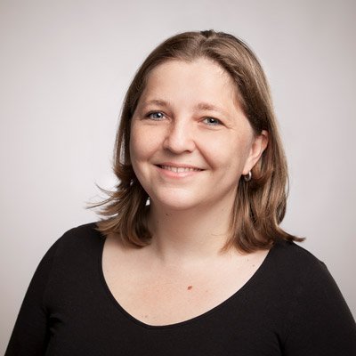 Eine lächelnde Frau mit mittellangen, braunen Haaren trägt ein schwarzes Oberteil und steht vor einem neutralen Hintergrund.