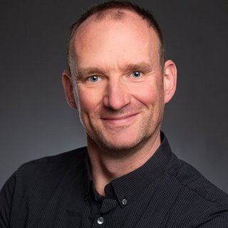 Professionelles Porträt eines selbstbewussten Mannes Ein lächelnder Mann mit kurzen Haaren trägt ein schwarzes Hemd und posiert vor einem neutralen Hintergrund.
