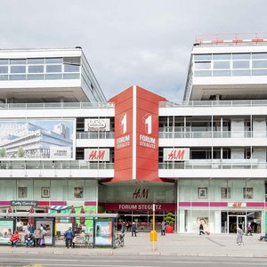 Forum Steglitz: Ein beliebter Ort zum Shoppen Vor dem Forum Steglitz stehen Menschen an einer Bushaltestelle, während das Gebäude mit Geschäften und Cafés belebt ist.