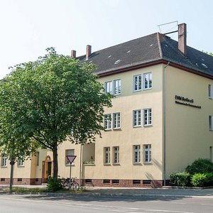 Städtische Architektur mit einladendem Straßenbild Die Fassade eines mehrstöckigen Gebäudes in sanften Gelbtönen, umgeben von üppigem Grün und einer Haltestelle für öffentliche Verkehrsmittel.