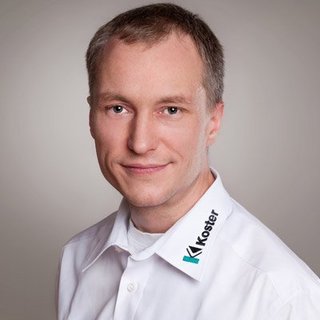 Professionelles Porträt eines lächelnden Mannes in Hemd Ein Mann mit kurzem, braunem Haar lächelt freundlich in die Kamera, trägt ein weißes Hemd mit einem Logo auf dem Kragen.