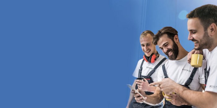 Drei Handwerker in Arbeitskleidung lachen und schauen auf ein Smartphone, während sie eine Tasse Kaffee halten.